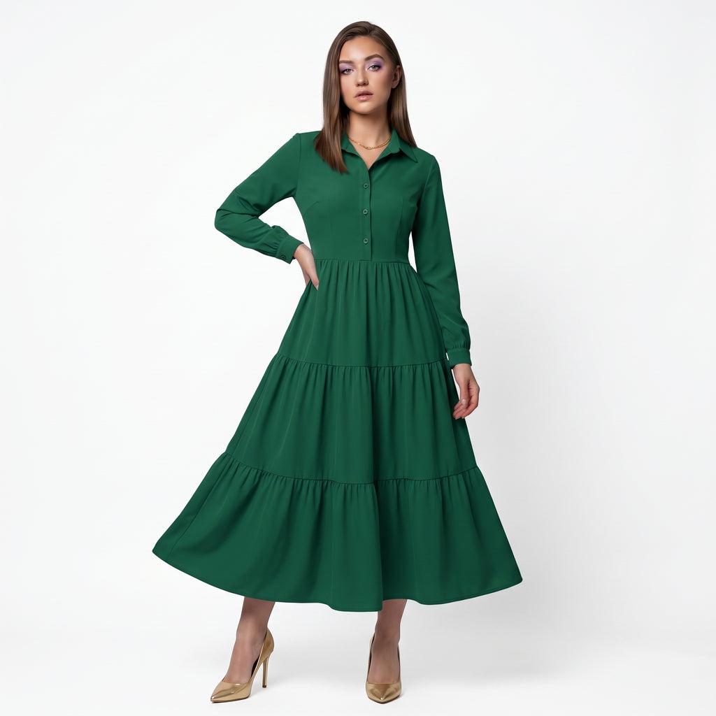 A woman modeling a long-sleeve green midi dress with a tiered skirt and buttoned collar, standing against a plain white studio background.