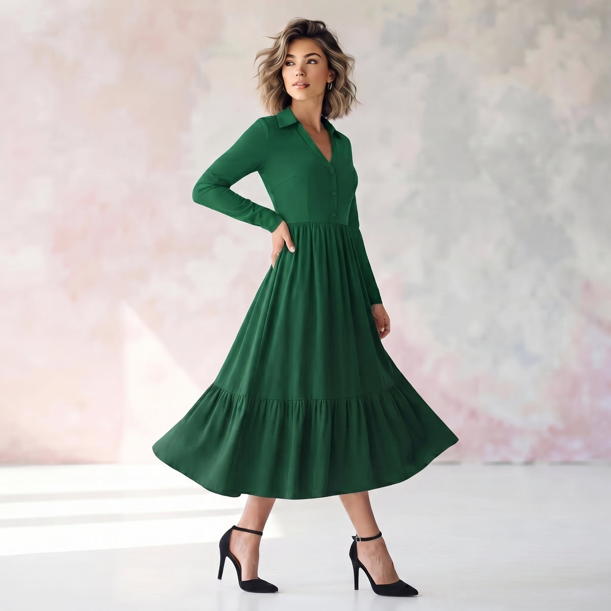 A woman modeling a long-sleeve green midi dress with a tiered skirt and black heels, standing against a soft pink and gray textured studio backdrop.