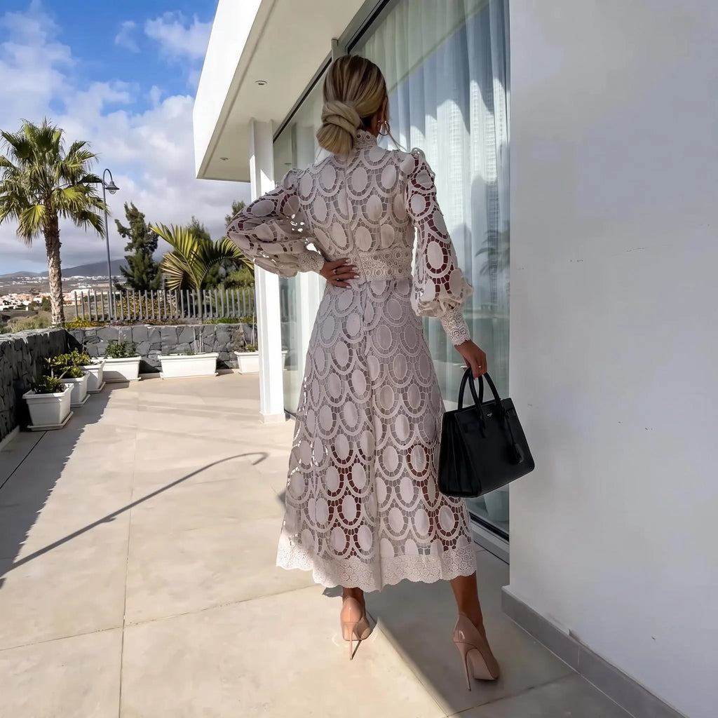 A woman stands on a sunlit outdoor terrace wearing a cream lace maxi dress with puff sleeves and a fitted waist, holding a black handbag. The dress has an intricate floral lace pattern.