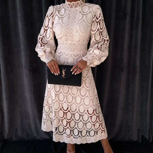 A woman wearing a cream lace midi dress with long balloon sleeves and a high neckline, holding a black clutch with a gold YSL logo, posed against a dark gray velvet curtain.