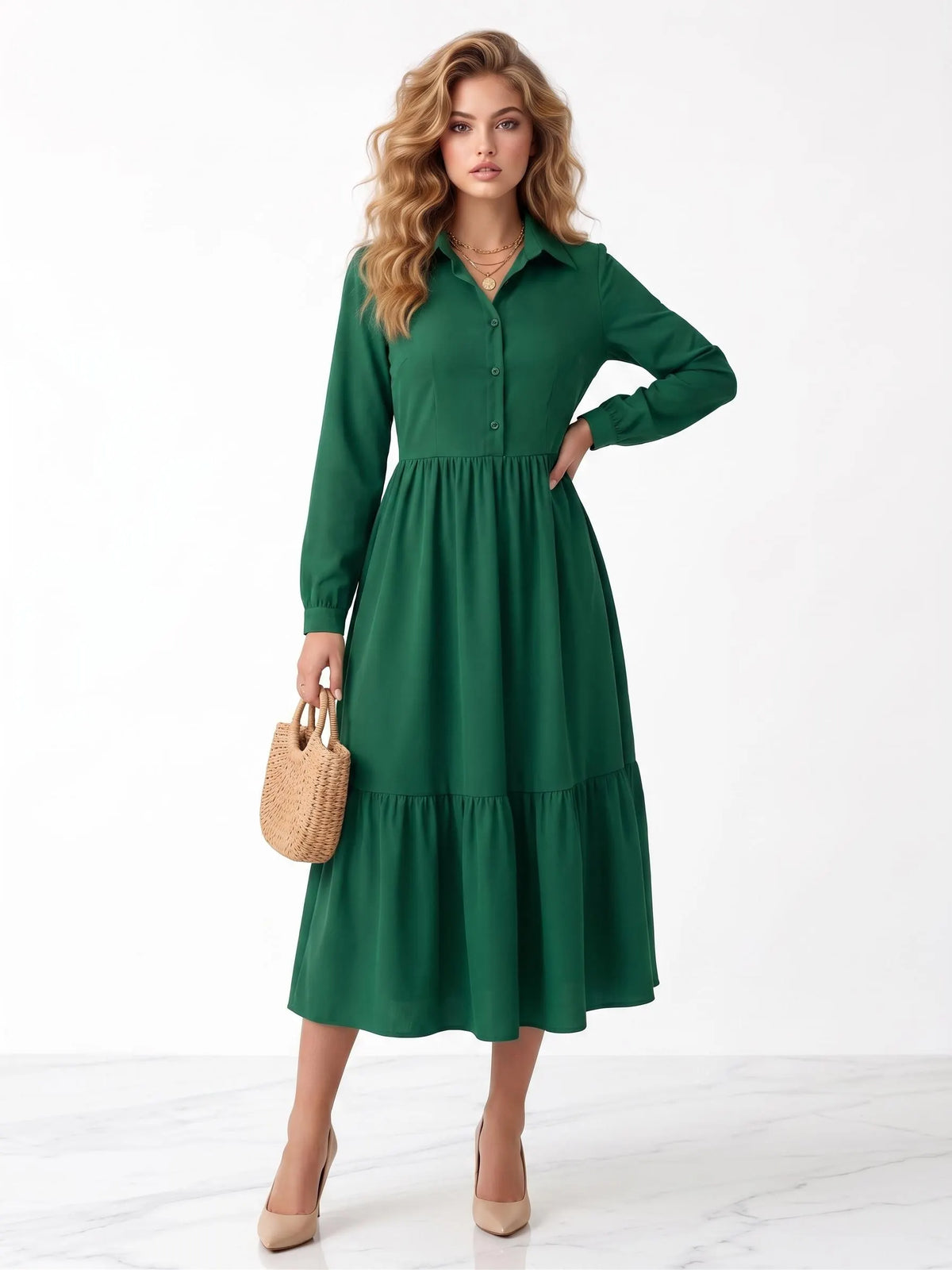 A woman modeling a long-sleeve green midi shirt dress with a tiered skirt, holding a small woven straw handbag and wearing beige heels against a clean white studio background.